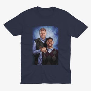 May include: Navy blue t-shirt featuring a printed image of two men wearing argyle vests over button-down shirts. The image has a blue background. The men are smiling and appear to be posing for a photo.