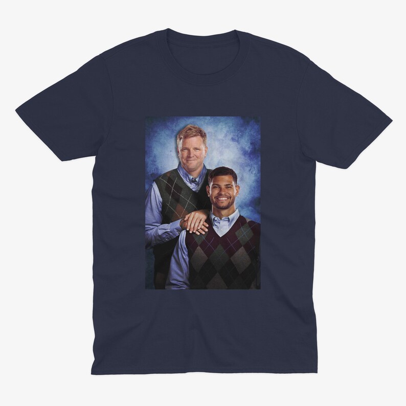 May include: Navy blue t-shirt featuring a printed image of two men wearing argyle vests over button-down shirts. The image has a blue background. The men are smiling and appear to be posing for a photo.