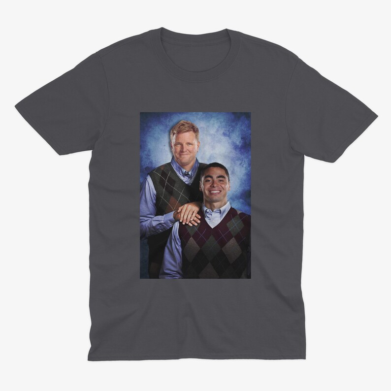 Peut inclure: Un t-shirt gris fonc&eacute; avec une photo de deux hommes portant des pulls. L'homme &agrave; gauche porte une chemise bleue et un pull &agrave; motifs vert et marron. L'homme &agrave; droite porte une chemise bleue et un pull &agrave; motifs rouge et blanc. Les hommes sont debout devant un fond bleu et blanc.