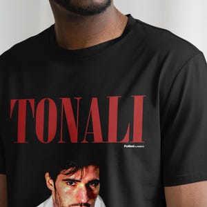以下が含まれることがあります： 黒のTシャツに赤い「TONALI」のグラフィックと男性の顔の写真がプリントされています。グラフィックの下に小さな文字で「Football Classics」と印刷されています。