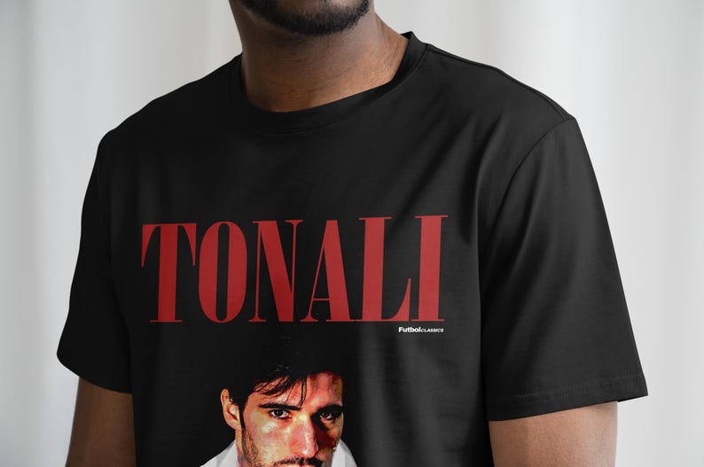 以下が含まれることがあります： 黒のTシャツに赤い「TONALI」のグラフィックと男性の顔の写真がプリントされています。グラフィックの下に小さな文字で「Football Classics」と印刷されています。