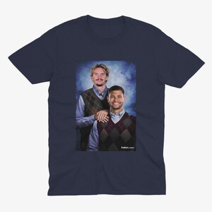 May include: Navy blue t-shirt featuring a portrait of two men wearing argyle vests over button-down shirts. The image has a blue background and the text "Futbol Classics" is visible.