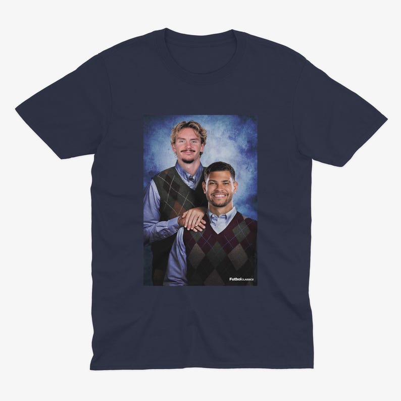 May include: Navy blue t-shirt featuring a portrait of two men wearing argyle vests over button-down shirts. The image has a blue background and the text "Futbol Classics" is visible.