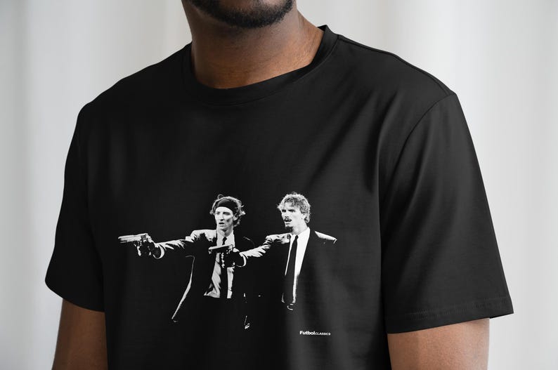 May include: Black t-shirt featuring a black and white graphic of two men in suits, each holding a gun. The image is a stylized representation of a scene from a film. The text "Futbol.Lances" is printed at the bottom.
