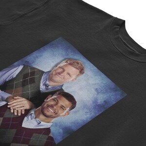 May include: Black t-shirt featuring a printed image of two people wearing argyle vests and button-down shirts. The image has a blue background. The t-shirt is made of a soft, comfortable material.
