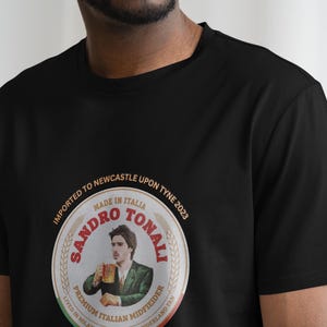Pu&ograve; includere: Maglietta nera con una grafica circolare di un viso maschile con il testo "SANDRO TONALI" in font rosso e oro. La grafica include anche il testo "IMPORTED TO NEWCASTLE UPON TYNE 2023" e "MADE IN ITALIA".