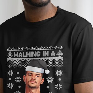 K&ouml;nnte beinhalten: Schwarzes T-Shirt mit festlichem Design. Das Shirt zeigt den Text "WALKING IN A" &uuml;ber einer Grafik eines Mannes mit Weihnachtsm&uuml;tze. Das Design umfasst Schneeflocken und einen Weihnachtsbaum.