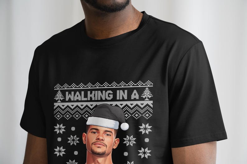 K&ouml;nnte beinhalten: Schwarzes T-Shirt mit festlichem Design. Das Shirt zeigt den Text "WALKING IN A" &uuml;ber einer Grafik eines Mannes mit Weihnachtsm&uuml;tze. Das Design umfasst Schneeflocken und einen Weihnachtsbaum.
