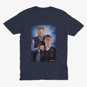 Op de afbeelding: Een marineblauw t-shirt met een foto van twee mannen die truien dragen. De man links draagt een blauw shirt en een bruin-witte geruite trui. De man rechts draagt een blauw shirt en een zwart-witte geruite trui. De tekst "Football Classics" is zichtbaar onderaan de afbeelding.