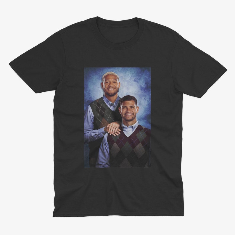 K&ouml;nnte beinhalten: Ein schwarzes T-Shirt mit einem Foto von zwei M&auml;nnern, die Pullover tragen. Die M&auml;nner stehen vor einem blauen und wei&szlig;en Hintergrund. Der Mann links tr&auml;gt ein blaues Hemd und einen gr&uuml;n-braunen Pullover. Der Mann rechts tr&auml;gt ein blaues Hemd und einen braun-wei&szlig;en Pullover.
