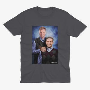 May include: A dark gray t-shirt with a photo of two men wearing sweaters. The man on the left is wearing a blue sweater with a brown vest and the man on the right is wearing a blue shirt with a brown and red patterned vest. The background is a blue and white gradient.