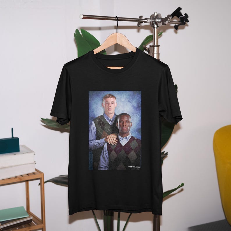 May include: Black t-shirt with a photo of two men wearing sweaters. The men are standing next to each other and smiling. The photo is set against a blue background.