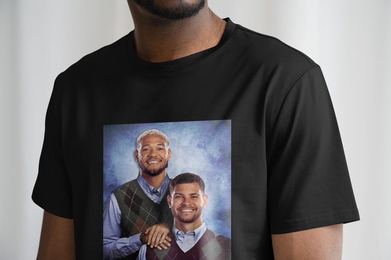 May include: Black t-shirt with a printed image of two men wearing sweaters with a patterned design. The men are smiling and standing next to each other.