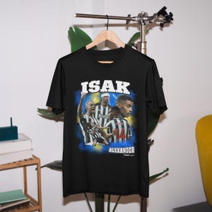 Puede incluir: Camiseta negra con un estampado gr&aacute;fico de un jugador de f&uacute;tbol, "ISAK ALEXANDER" en texto blanco y un fondo azul y amarillo.