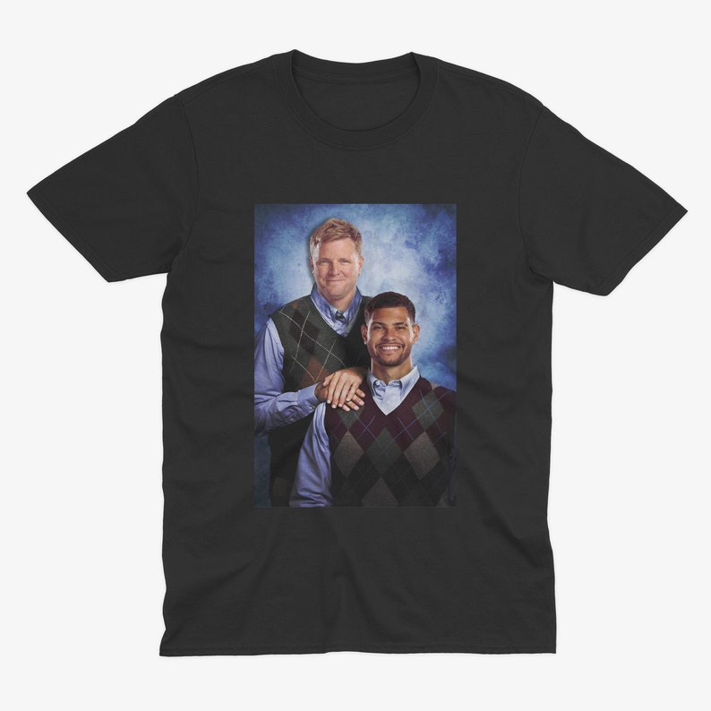 May include: Black t-shirt featuring a printed image of two men. The men are wearing argyle vests over button-down shirts. One man has his arm on the other's shoulder. The background is a faded blue.