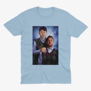 Op de afbeelding: Een lichtblauw t-shirt met een foto van twee mannen die truien dragen. De man links draagt een groen-bruine trui met een patroon en de man rechts draagt een bruin-witte trui met een patroon.