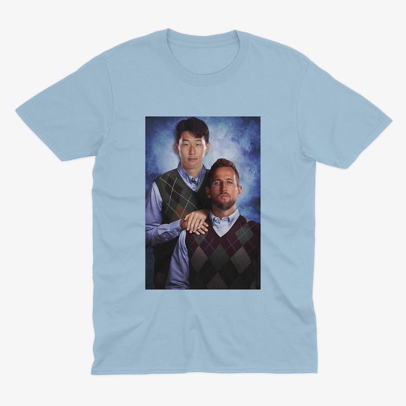 Op de afbeelding: Een lichtblauw t-shirt met een foto van twee mannen die truien dragen. De man links draagt een groen-bruine trui met een patroon en de man rechts draagt een bruin-witte trui met een patroon.