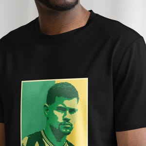 K&ouml;nnte beinhalten: Schwarzes T-Shirt mit einem Portr&auml;t auf der Vorderseite. Das Portr&auml;t hat einen gr&uuml;nen und gelben Hintergrund und zeigt einen Mann mit Bart. Das T-Shirt ist ein Rundhals-Modell.