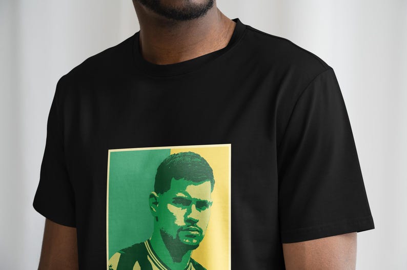 K&ouml;nnte beinhalten: Schwarzes T-Shirt mit einem Portr&auml;t auf der Vorderseite. Das Portr&auml;t hat einen gr&uuml;nen und gelben Hintergrund und zeigt einen Mann mit Bart. Das T-Shirt ist ein Rundhals-Modell.