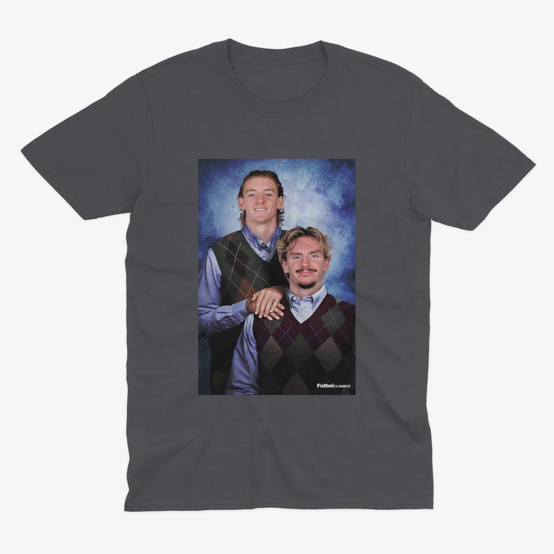 May include: Dark gray t-shirt featuring a portrait of two people wearing argyle vests over button-down shirts. The image has a blue background and the text "Futbol Classics" is visible.