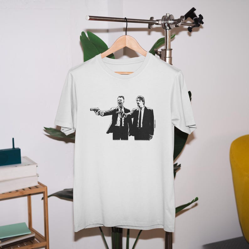 May include: White t-shirt featuring a black and white graphic of two men in suits, one pointing a gun. The image is a stylized illustration. The shirt is hanging on a wooden hanger.