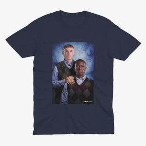 May include: A navy blue t-shirt featuring a photo of two men in sweaters. The men are standing next to each other and smiling. The background is a blue and white gradient.
