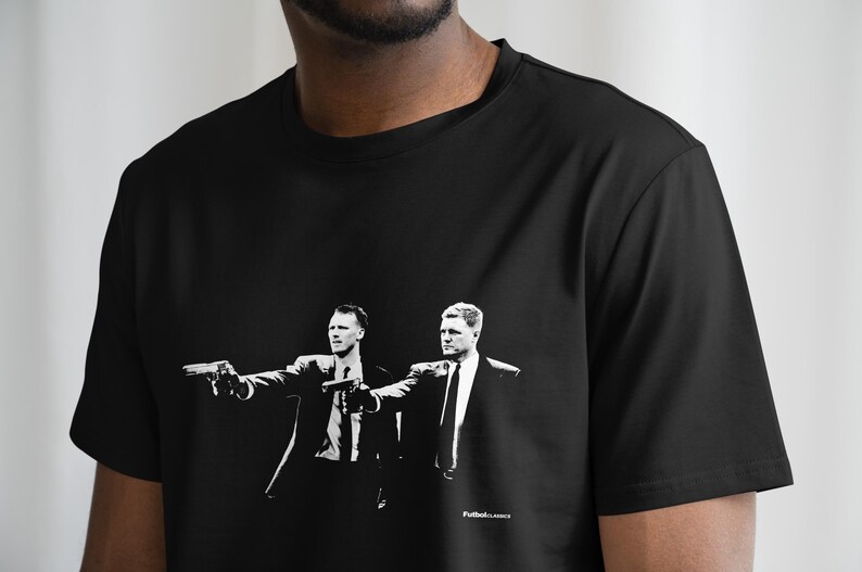 May include: Black t-shirt featuring a black and white graphic of two men in suits holding guns. The image is a stylized illustration. The text "Futbol.classics" is printed in white on the bottom right of the shirt.