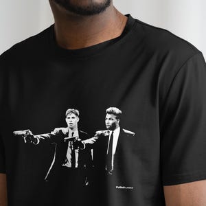 Op de afbeelding: Zwart T-shirt met een witte grafische print van twee mannen in pak die wapens vasthouden. De tekst "Pulp Fiction" is onderaan de print gedrukt.