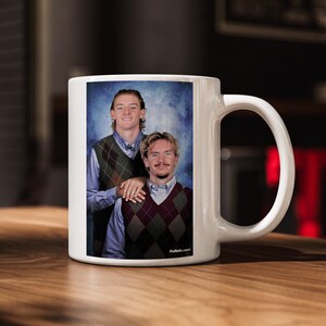 May include: White ceramic mug featuring a portrait of two men in argyle vests and collared shirts. The image has a vintage aesthetic. The mug has a large handle and is set on a wooden surface.