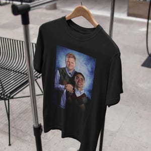 Peut inclure: T-shirt noir avec une image imprim&eacute;e de deux hommes en pulls sur un fond bleu. Les hommes sourient et regardent la cam&eacute;ra.