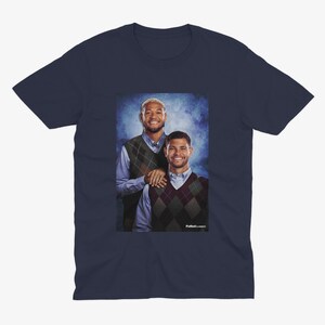 May include: A navy blue t-shirt featuring a photo of two men wearing sweaters. The man on the left is wearing a green sweater vest and the man on the right is wearing a brown and white argyle sweater vest. The men are standing in front of a blue and white background.