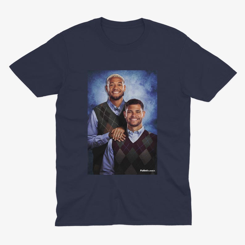 May include: A navy blue t-shirt featuring a photo of two men wearing sweaters. The man on the left is wearing a green sweater vest and the man on the right is wearing a brown and white argyle sweater vest. The men are standing in front of a blue and white background.