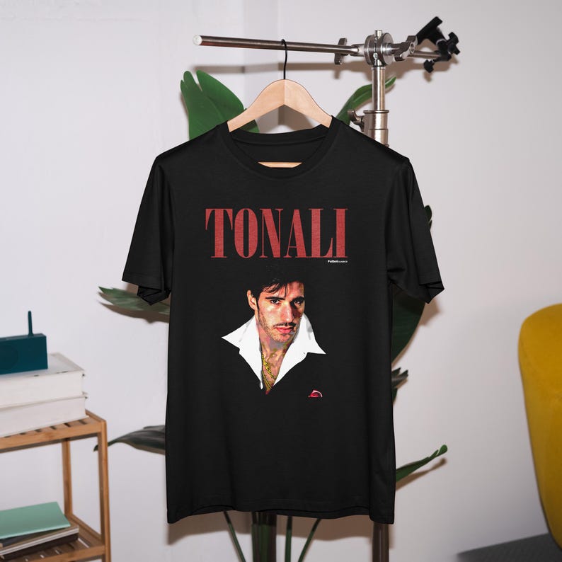 以下が含まれることがあります： 黒いTシャツに赤い「TONALI」の文字と、白い襟付きシャツを着た黒髪の男性の写真がプリントされています。