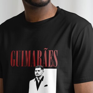 May include: Black t-shirt featuring the name "GUIMAR&Atilde;ES" in red, above a black and white portrait of a man in a suit. The shirt is made of a soft material.