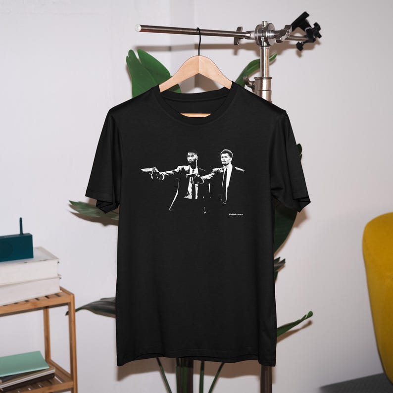 Op de afbeelding: Zwart T-shirt met een witte afbeelding van twee mannen in pak, een van hen wijst met een pistool, met de tekst "Pulp Fiction" eronder.