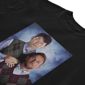 Op de afbeelding: Een zwart sweatshirt met een bedrukte afbeelding van twee mannen die geruite vesten dragen. De mannen lachen en kijken naar de camera. De achtergrond is een blauwe en witte gradi&euml;nt.
