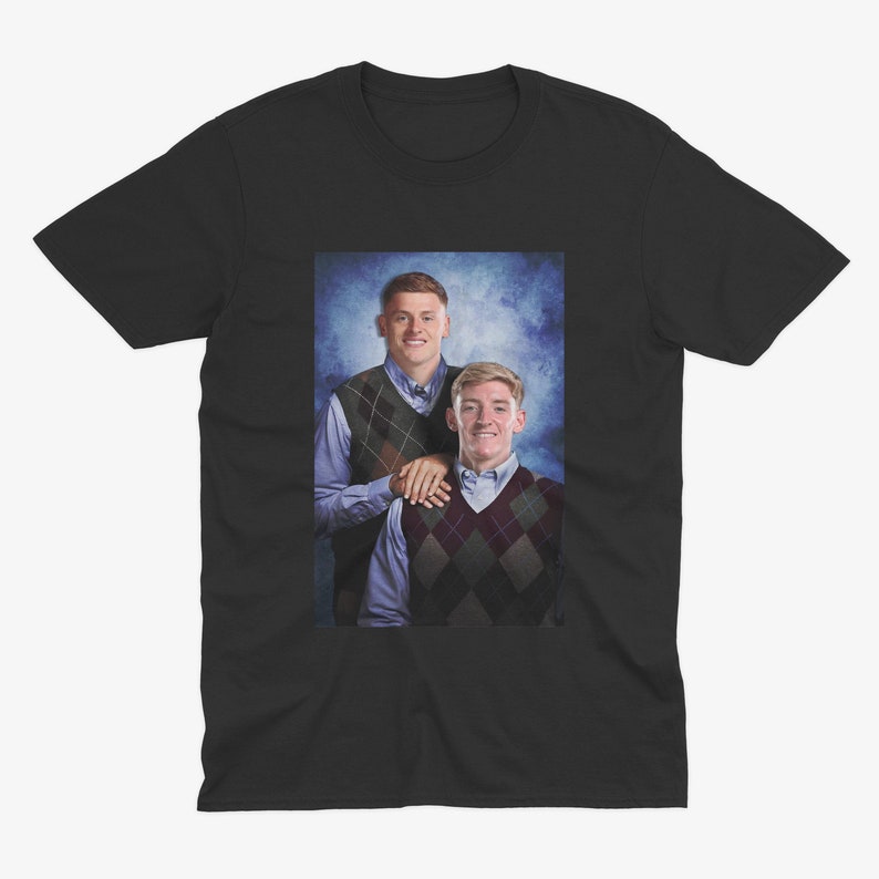 May include: A black t-shirt featuring a photograph of two men wearing sweaters. The men are standing side-by-side and smiling. The background is a blue and white gradient.