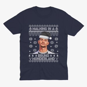 K&ouml;nnte beinhalten: Marineblaues T-Shirt mit weihnachtlichem Design. Das Shirt zeigt ein Portr&auml;t einer Person mit Weihnachtsm&uuml;tze, mit dem Text "WALKING IN A" dar&uuml;ber und "BRUNO WONDERLAND" darunter, umgeben von Schneeflocken und Weihnachtsb&auml;umen.