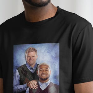 May include: A black t-shirt with a printed image of two men wearing sweaters. The men are smiling and standing next to each other. The background is a blue and white gradient.