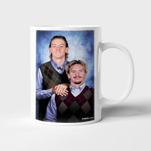 May include: White ceramic mug featuring a portrait of two people wearing argyle vests over button-down shirts. The image has a blue background and the text "FutbolClassics" is printed on the mug.