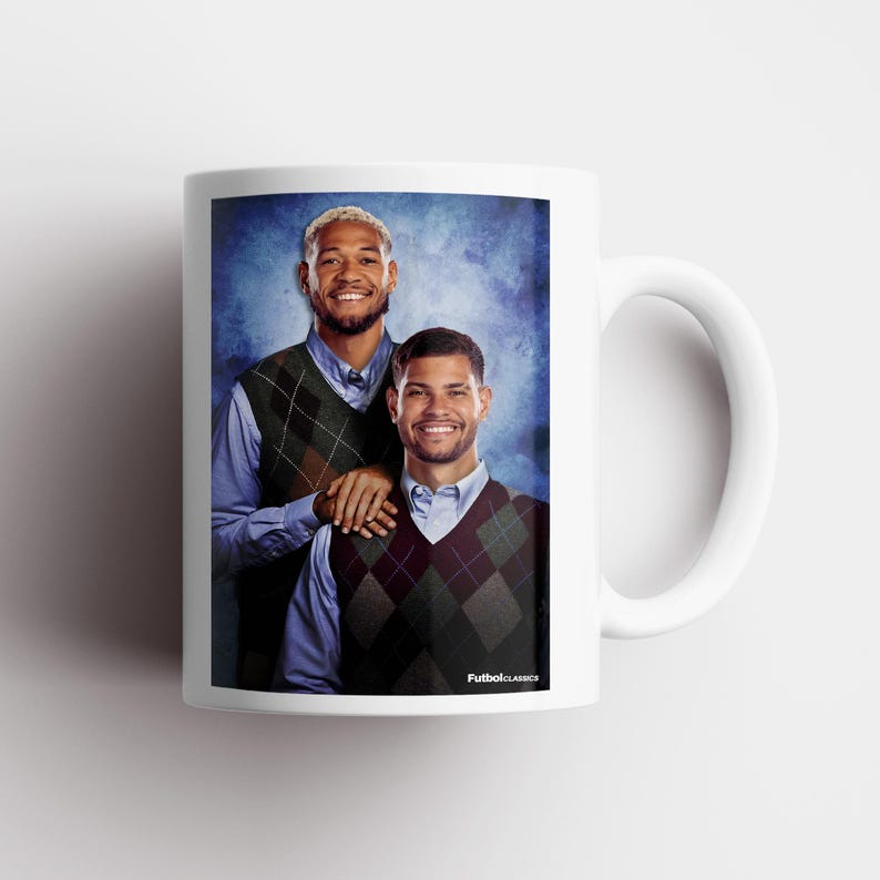 May include: White ceramic mug featuring a portrait of two smiling men wearing argyle vests over blue shirts. The mug has a white handle and the text "FutbolClassics" is printed on the side.