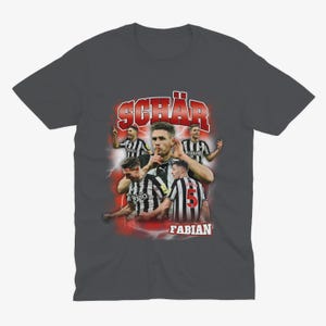 May include: A dark grey t-shirt featuring a graphic print of a football player, Fabian Sch&auml;r, in a black and white kit. The graphic has a retro style with red and white accents.