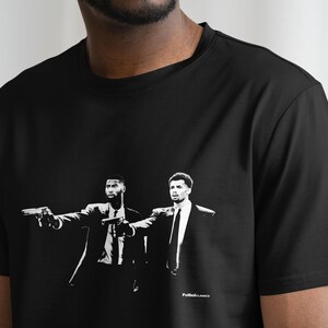 Op de afbeelding: Zwart-wit T-shirt met een grafische print van twee mannen in pak die met pistolen naar de kijker wijzen. De tekst "Pulp Fiction" staat onderaan het shirt.