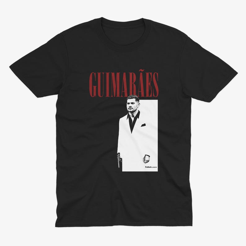 May include: Black t-shirt featuring a graphic of a man in a white suit with the name "GUIMAR&Atilde;ES" in red above the image. The design is a stylized illustration, likely a tribute or fan art.