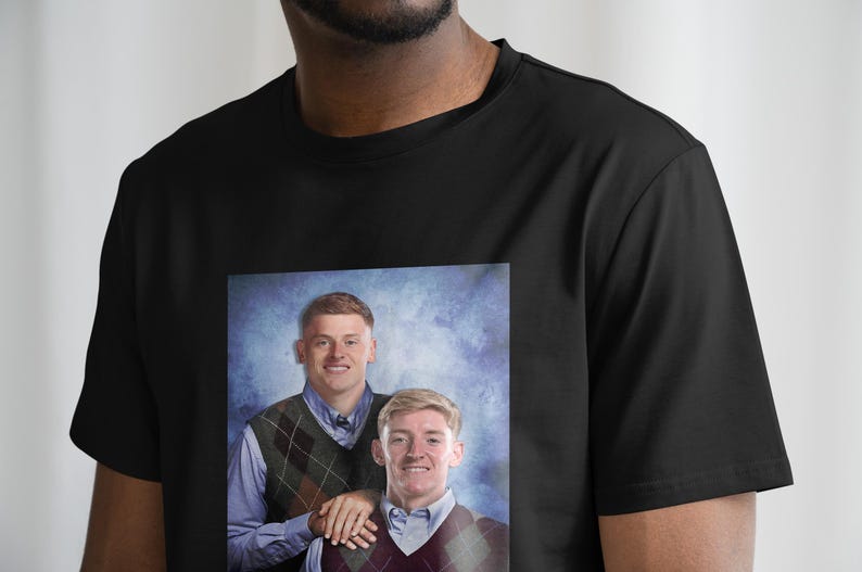 May include: A black t-shirt featuring a printed image of two men wearing sweaters. The men are standing in front of a blue and white backdrop. The image is a portrait style photograph.