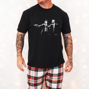 May include: Black t-shirt featuring a black and white graphic of two men in suits pointing guns. The person in the image is wearing red, white, and black plaid pajama pants. The shirt is a crew neck.