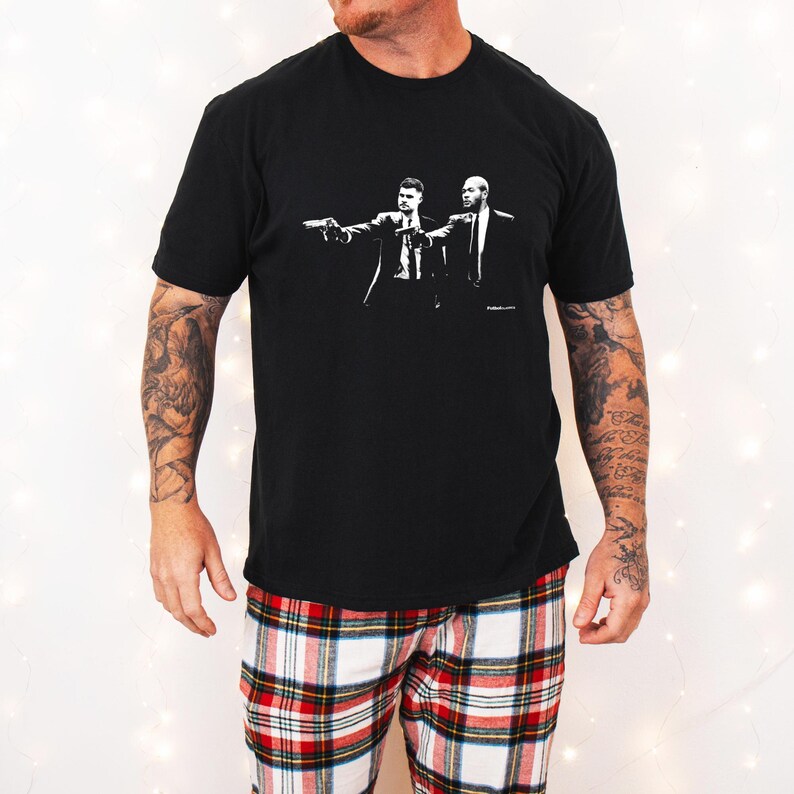 May include: Black t-shirt featuring a black and white graphic of two men in suits pointing guns. The person in the image is wearing red, white, and black plaid pajama pants. The shirt is a crew neck.