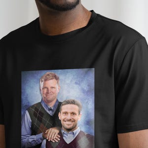May include: A black t-shirt with a printed image of two men wearing sweaters. The men are smiling and standing next to each other. The background is a blue and white gradient.