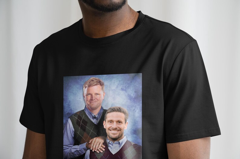 May include: A black t-shirt with a printed image of two men wearing sweaters. The men are smiling and standing next to each other. The background is a blue and white gradient.