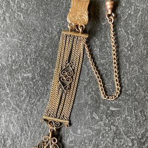 May include: Antique gold-toned watch chain with a decorative rectangular top and a series of small chains. The chain features an ornate design with a heart-shaped element and a decorative seal at the bottom. A small chain with a clasp is attached.
